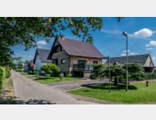 Wohngebiet mit Einfamilienhäusern; im Vordergrund ein Haus mit spitzem Dach, heller Fassade und Balkon, umgeben von einem gepflegten Garten mit Rasenflächen und Zierbäumen. Rechts neben dem Haus ein Gehweg, im Hintergrund weitere ähnliche Häuser und blauer Himmel.