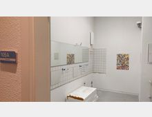 Innenraum eines einfachen Waschraums mit weißen Fliesen an den Wänden und einem großen Spiegel. Die Einrichtung ist unvollständig, mit unbenutzten Armaturen und Beschädigungen an der Wand; an der Tür ein Schild mit der Aufschrift „105A Zahnputzraum“.