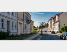 Wohnstraße in einem Wohnviertel mit mehrgeschossigen Altbauten und modernen Wohnhäusern, teils mit roten Ziegeldächern. Eine asphaltierte Straße mit Gehweg, Kopfsteinpflasterrand, jungen Straßenbäumen und parkenden Autos führt in eine leichte Kurve.
