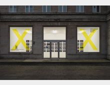 Fassade einer Architekturgalerie in Berlin mit zwei großen Schaufenstern, auf denen jeweils ein gelbes „X“ abgebildet ist. Im Inneren sind beleuchtete Ausstellungswände mit zahlreichen Bildern oder Dokumenten zu sehen.