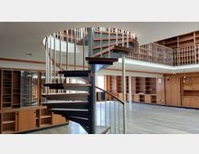 Offener Innenraum mit einer freistehenden Wendeltreppe aus Holz und Metall in der Mitte. Der Raum ist von hölzernen Regalen und Einbauschränken umgeben, mit einem offenen Zwischengeschoss, das von weißen Geländern begrenzt wird.
