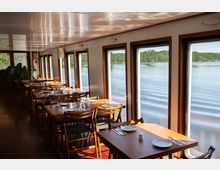 Innenraum eines Restaurants auf einem Boot mit Holzböden und -möbeln. Große Fenster auf der rechten Seite bieten einen Ausblick auf ein ruhiges Gewässer und grüne Uferlandschaft.