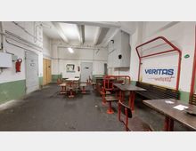 Ein einfacher Aufenthaltsraum in einem industriellen Gebäude mit mehreren rot-weißen Tischen und Sitzbänken, weißen Wänden mit grüner Sockelbemalung und sichtbaren Versorgungsleitungen. Im Hintergrund befinden sich Türen, ein Feuerlöscher und eine Beschriftung mit dem Text „VERITAS weltweit“.