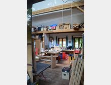 Innenraum einer Holzwerkstatt mit einem Arbeitsbereich, der von Holzbänken, Werkzeugen und Materialien bedeckt ist. Im Hintergrund befinden sich Fenster mit Vorhängen, eine hohe Decke mit sichtbaren Holzbalken und ein Regal mit gelagerten Holzstücken und Kisten.