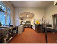 Großküche mit roten Fliesenboden, weißen Wänden mit gefliesten Bereichen und Edelstahl-Arbeitsflächen. Der Raum enthält Spülbecken, eine Industriespülmaschine, Regale mit Geschirr und Kochutensilien sowie mehrere Fenster, die Tageslicht hereinlassen.