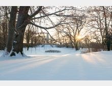 Schneebedeckter Park mit großen, kahlen Bäumen im Vordergrund. Im Hintergrund ist ein kleines Gebäude mit Säulen und Treppen zu sehen, während die tiefstehende Sonne durch die Äste scheint.