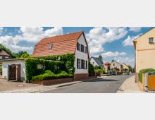 Straßenszene in einem Wohnviertel mit Einfamilienhäusern, darunter ein weißes Haus mit rotem Satteldach und Efeu an der Fassade. Die Straße ist asphaltiert, gesäumt von schmalen Gehwegen und vereinzelten Bäumen, bei klarem Himmel mit Wolken.