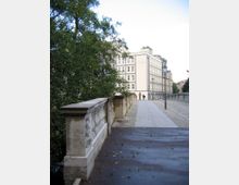 Steinbrücke mit verziertem Geländer, flankiert von Bäumen auf der linken Seite und einem mehrstöckigen Gebäude im Hintergrund. Der gepflasterte Weg über die Brücke führt zu einer hellen Fassade mit Fenstern und lässt zwei Personen in der Ferne erkennen.