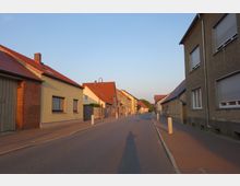 Straßenansicht in einem Dorf mit dicht aneinander gebauten Wohnhäusern, die überwiegend verputzt sind und rote Ziegeldächer haben. Ein schmaler Gehweg mit Pollern verläuft entlang der Straße, und die Szene wird von einem klaren Himmel bei Sonnenuntergang beleuchtet.