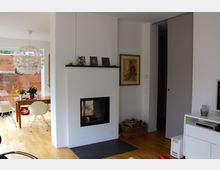 Helles Wohnzimmer mit einem modernen Kamin in einer weißen Wand. Im Hintergrund ein Essbereich mit Holztisch und weißen Stühlen, daneben eine große Fensterfront mit Blick auf eine Holzverkleidung im Außenbereich.