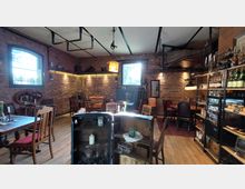 Gemütliches Café oder Restaurant im rustikalen Stil mit Wänden aus rotem Ziegelstein, Holzmöbeln und zahlreichen dekorativen Elementen wie Regalen mit Getränken und Kerzen. Der Raum ist mit warmem Licht beleuchtet, hat große Fenster und eine Mischung aus Tischen, Stühlen und Sesseln.