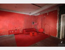 Ein Raum mit abblätterndem roten Putz an den Wänden, einer roten Ledercouch, einem roten Teppich und roten Möbelstücken, darunter ein Beistelltisch, ein Sideboard, ein Sessel und zwei Lampen. Die Decke zeigt unverkleidete Beton-Elemente, und der Boden besteht aus einer Betonfläche.