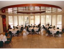 Ein heller Veranstaltungsraum mit mehreren runden Tischen, die mit weißen Tischdecken, Geschirr und Dekoration gedeckt sind. Große Fenster mit halbtransparenten Vorhängen lassen Tageslicht herein, und die Decke ist mit Holzringen und integrierter Beleuchtung verziert.