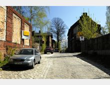 Straßenszene mit Kopfsteinpflaster in einer ländlichen Umgebung, gesäumt von alten Backsteinbauten mit teils verwitterten Fassaden. Mehrere parkende Autos und frische Baumtriebe sind sichtbar, im Hintergrund stehen größere Bäume unter klarem Himmel.