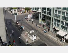 Straßenszene am Checkpoint Charlie in Berlin, mit einer Nachbildung eines historischen Kontrollhäuschens in der Mitte der Straße. Im Hintergrund sind ein Poster mit einem Soldatenporträt, mehrere Passanten und ein modernes Gebäude mit dem Schriftzug „Wall Museum“ sichtbar.