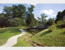 Eine Parklandschaft mit einem geschwungenen gepflasterten Weg, der zu einer filigranen Metallbrücke über einen kleinen Bach führt. Umgeben von sanften Grünflächen, Bäumen und Büschen unter einem klaren Himmel.