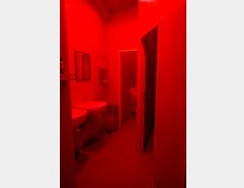 Ein Badezimmer mit roter Beleuchtung, ausgestattet mit zwei Waschbecken, einem Mülleimer mit Drahtkorb und einer offenen Tür, die zu einer Toilette führt.