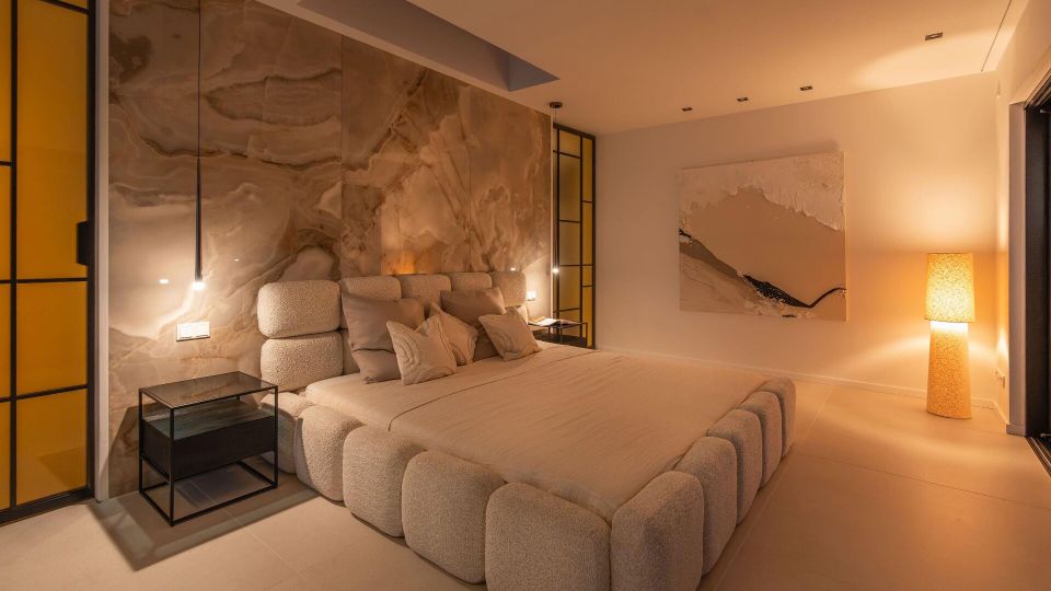 Modernes Schlafzimmer mit großem gepolstertem Bett und heller Bettwäsche vor einer großformatigen Marmor-Akzentwand. Warmes indirektes Licht, schmale Nachttische, gelb getönte Glas-Elemente und ein großes abstraktes Wandbild sowie eine Stehlampe prägen den minimalistischen Raum.
