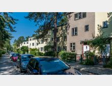 Reihenhäuser mit flachen Dächern und hellen Fassaden entlang einer von Bäumen gesäumten, gepflasterten Straße mit parkenden Autos. Der Vorgartenbereich ist durch Zäune und Grünflächen begrenzt, im Hintergrund sind weitere Bäume sichtbar.