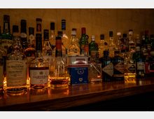 Eine Barablage mit einer Vielzahl von Alkoholflaschen, darunter Whiskey, Cognac und Gin, in verschiedenen Formen und Farben, vor einer schlichten, goldbraunen Ziegelwand. Die Beleuchtung hebt die Labels und Flaschen hervor.