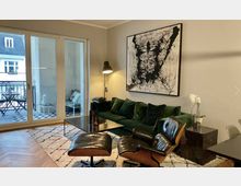 Modernes Wohnzimmer mit grünem Samtsofa, schwarzem Ledersessel mit Fußhocker und abstraktem Kunstwerk an der Wand. Große Fensterfront mit Zugang zu einem kleinen Balkon, auf dem sich ein Tisch und Stühle befinden, sowie helle Wände und ein gemusterter Teppich.