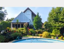 Zweistöckiges Einfamilienhaus mit weißer Fassade und rotem Satteldach, umgeben von einem gepflegten Garten mit Blumen, Büschen und Bäumen. Im Vordergrund ein rundes Schwimmbecken mit Einstiegstreppe, das von einer gepflasterten Terrasse umgeben ist.