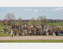 Eine städtische Gemeinschaftsgartenszene mit zahlreichen Hochbeeten, improvisierten Strukturen und bunten Dekorationen, umgeben von einer Grünfläche und Bäumen. Im Hintergrund sind Wiesen und eine Reihe von Gebäuden am Horizont zu sehen.