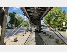 Ein Blick unter eine Hochbahntrasse in einer urbanen Umgebung mit einer Straße auf beiden Seiten. Unter der Trasse befinden sich ein Geh- und Radweg, Autos, sowie große Steinblöcke; im Hintergrund sind Bäume, Gebäude und Verkehrsschilder sichtbar.