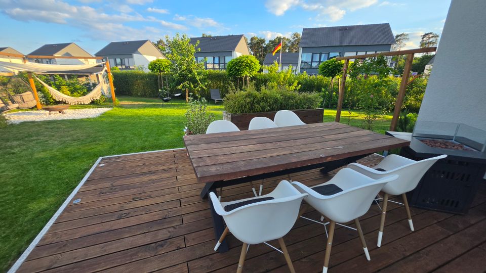 Holzterrasse mit großem rechteckigem Esstisch und mehreren weißen Stühlen, neben einer Außenfeuerstelle. Dahinter ein gepflegter Garten mit Rasen, Hecken, Hochbeet und Hängematte unter einem Sonnensegel; im Hintergrund stehen moderne Wohnhäuser.