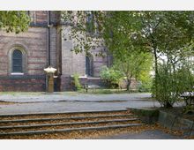 Ein Außenbereich mit der Backsteinfassade einer historischen Kirche. Zu sehen sind Spitzbogenfenster mit Gitterverglasung, eine goldfarbene Skulptur sowie eine kleine Grünanlage mit Bäumen und Bänken; im Vordergrund führen Stufen durch eine mit Herbstlaub bedeckte Fläche.