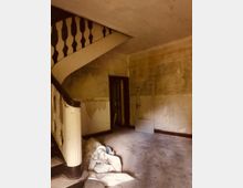 Eingangsbereich eines alten, renovierungsbedürftigen Gebäudes mit einer Holztreppe, abgenutzten Wänden und einer losen Tür, die an die Wand lehnt. Auf dem Boden liegen Baustellenmaterialien und Schmutz.