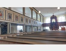 Innenansicht einer historischen Kirche mit Holzbänken, einer verzierten Empore mit Wappenmalereien und einem reich dekorierten barocken Altar. Große, rechteckige Fenster lassen Tageslicht in den hellen Raum.
