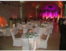 Ein festlich dekorierter Veranstaltungsraum in einem historischen Gewölbe mit Backsteinwänden. Rundtische mit weißen Tischdecken und Stuhlhussen sind gleichmäßig verteilt, dazu florale Dekorationen und Kerzenleuchter; im Hintergrund bunte Beleuchtung und eine Bühne mit Musikinstrumenten.