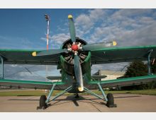 Ein grünes Doppeldecker-Flugzeug steht auf einem Flugfeld, frontal aufgenommen, mit sichtbarem Propeller und gestreckten Tragflächen. Im Hintergrund befinden sich ein Gebäude, ein hoher Mast mit Beleuchtung und Bäume, unter blauem Himmel mit wenigen Wolken.