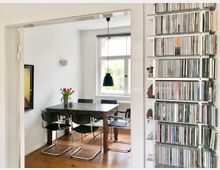 Esszimmer mit Holzboden und einem rechteckigen Esstisch aus dunklem Holz, umgeben von vier schwarzen Stühlen mit Chromgestellen. Im Hintergrund ein großes Fenster, daneben eine Wand mit Regalen voll CDs; auf dem Tisch steht eine Vase mit roten Tulpen.