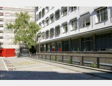 Ein moderner Gebäudekomplex mit weißer Fassade und zahlreichen Fenstern, teils mit Jalousien bedeckt. Im Vordergrund befinden sich eine gepflasterte Fläche, markierte Parkplätze und ein einzelner Baum; ein Gerüst und ein roter Bauzaun sind im Hintergrund sichtbar.