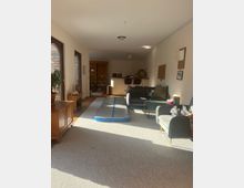 Wohnraum mit hellen Wänden, Teppichboden und großen Fenstern mit Holzrahmen. Im Raum befinden sich eine Luftmatte, ein Sofa, Sessel, ein Holztisch sowie ein Regal, und im Hintergrund ist eine offene Küchenecke zu sehen.