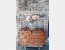 Eine alte Ziegelwand mit sichtbaren Gebrauchsspuren, vor der eine rustikale Holztruhe auf Rollen steht. Auf der Truhe befindet sich eine gläserne Vase mit rosa Pfingstrosen.