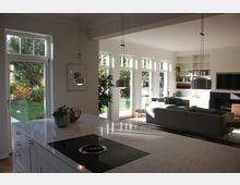 Moderner, offener Wohnbereich mit Kücheninsel im Vordergrund, einem grauen Sofa und einem Kamin im Hintergrund. Große Fenster lassen viel Tageslicht herein und bieten einen Blick auf den Garten.