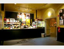 Kleiner Verkaufsbereich in einem Kino mit Theke, auf der Snacks, Getränke und eine Popcornmaschine stehen. Im Hintergrund befinden sich Filmposter an der Wand, daneben eine offene Tür mit Blick auf einen Raum mit Backsteinwand und Dekoration.