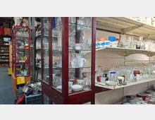 Innenansicht eines Trödelladens mit hohen Glasvitrinen und weißen Wandregalen, dicht gefüllt mit Gläsern, Tassen, Schalen und Geschirr. Zwischen den Regalen stehen Kisten und gestapelte Haushaltswaren, wodurch ein enger, vollgestellter Gang entsteht.