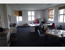 Ein einfacher Büroraum mit mehreren Schreibtischen, Bürostühlen und Regalen. Die Wände sind weiß, die Fenster haben Jalousien, und der Boden ist mit dunkelgrauem Teppich ausgelegt.