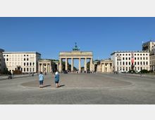 Das Bild zeigt das Brandenburger Tor in Berlin, ein neoklassizistisches Bauwerk mit sechs Säulen und einer Quadriga auf der Spitze. Links und rechts des Tors befinden sich symmetrische Seitengebäude, umgeben von modernen Verwaltungsgebäuden; im Vordergrund ist ein großer gepflasterter Platz mit Besuchern.