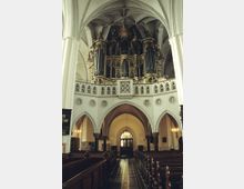 Innenraum einer historischen Kirche mit hohen weißen Bögen und Gewölbedecken. Im Vordergrund befinden sich Holzbänke, im Hintergrund eine reich verzierte Orgel auf einer Empore über einem Durchgang.
