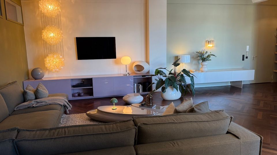 Modern living room with sectional sofas facing a wall-mounted TV above low built-in cabinetry, lit by a large drum ceiling light and cascading pendant chandelier. A sculptural coffee table, potted plants, and warm wall sconces sit over dark herringbone wood flooring with an open entry area to the right.