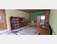 Wohnzimmer mit gelb- und grün gestrichenen Wänden, einer hölzernen Eingangstür und einem hellen Teppichboden. Das Zimmer enthält zwei Bücherregale, eine kleine rote Tischlampe, eine Anbauwand, eine rote Couch und einen Holztisch.