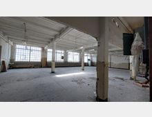 Ein leerer, verfallener Industriebereich mit hohen Decken, freiliegenden Stahlträgern und großen Fenstern mit Glasbausteinen. Der Raum wirkt verlassen mit Schutt auf dem Boden und bröckelnden Wänden.