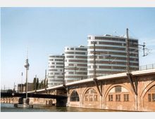 Moderne Bürogebäude mit abgerundeten Glasfassaden entlang eines eisenbahnführenden Viadukts aus rotem Backstein. Im Vordergrund fließt ein Fluss, und links ist der Berliner Fernsehturm sichtbar.
