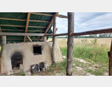 Ein Lehmofen unter einem schlichten Holzdach mit grünem Planenbezug, umgeben von einer ländlichen Wiesenlandschaft mit Feldern und Bäumen am Horizont.