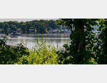 Blick auf einen See, eingefasst von dichtem, grünem Uferbewuchs, durch den Boote und Häuser am gegenüberliegenden Ufer sichtbar sind. Im Hintergrund erstreckt sich eine Baumreihe unter einem klaren Himmel.